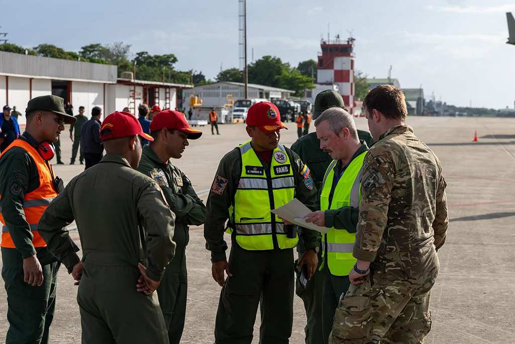 A group of five airport officials stand in a circle with two RAF Voyager personnel on the aircraft operating platform under a partly cloudy sky, as they discus details of the RAF Voyager. there are airport buildings in the background, including a red and white control tower.